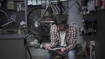 Tired bike shop owner fills out paperwork and does calculations for IRS, gets bored and drinks coffee. Bicycle mechanic drinks coffee with tablet in hands, calculating cost of repairing bicycle. video