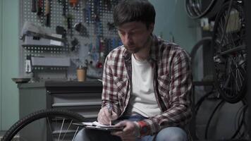 Male bicycle mechanic calculates cost of repairing a cycle with pen and clipboard in hand and yawns wearily and drinks coffee in bike shop. Using tablet while fixing bicycle. Tired repairman at work video