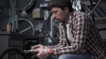 Tired bicycle mechanic drinks coffee at his workplace while calculating cost of working in bike shop. Overtime work. Boring paperwork. Inventorying items. Man calculates price of repairing the bike. video
