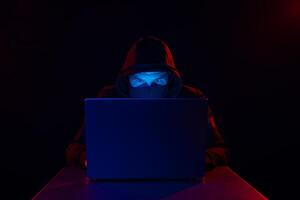 Cyber hacker in hood behind laptop on dark background with red blue light, cyber security concept photo