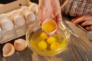 A person cracking an egg into a glass bowl, with several eggs in an egg carton in the background photo