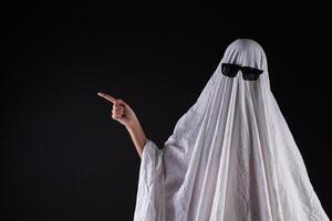 Person in costume of ghost pointing at something on dark background with space for tex photo