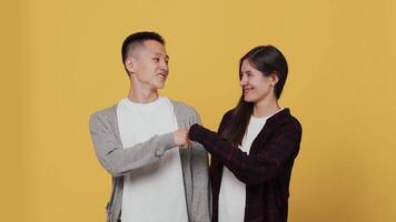 Young asian guy and a young caucasian girl standing next to each other, laughing and making a fist bump gesture enjoying collaboration on a yellow background in the studio video