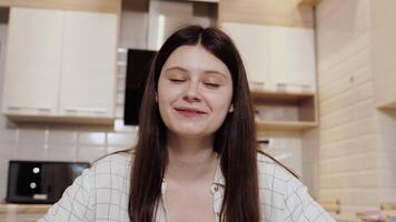 Front view of a young caucasian girl in casual clothes sitting at home in a cozy kitchen and having a pleasant conversation with her interlocutor via call video
