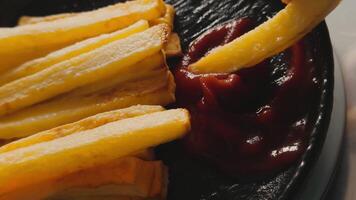 Piece of delicious french fries is dipped into the ketchup from a bundle, lying on a plate close up. A pile of appetizing fries with a sauce video