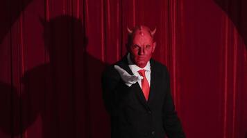 Red devil character wearing a business suit showing with a gesture that he can't hear you and waiting for your action standing on a stage with red curtains background under a spotlight video