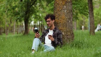 Young man relaxing under tree browsing smartphone and drinking coffee video