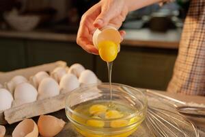 A person cracking an egg into a glass bowl, with several eggs in an egg carton in the background photo