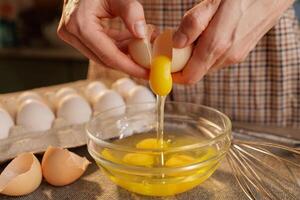 A person cracking an egg into a glass bowl, with several eggs in an egg carton in the background photo