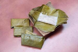 Steamed Banana Cake on a banana leaf. Placed on a plate with a coconut palm pattern. Wrapped banana cakes are lined up on the side. Placed on the red tile floor. photo