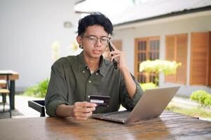 Young Asian man working on his laptop while talking on mobile phone and holding a credit card. Man trying to online purchasing with card photo