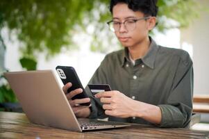 Young Asian man working on his laptop while talking on mobile phone and holding a credit card. Man trying to online purchasing with card photo