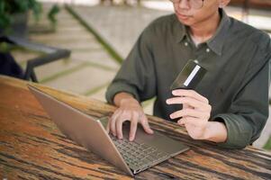 Young Asian man working on his laptop while talking on mobile phone and holding a credit card. Man trying to online purchasing with card photo