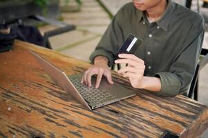 Young Asian man working on his laptop while talking on mobile phone and holding a credit card. Man trying to online purchasing with card photo