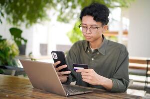 Young Asian man working on his laptop while talking on mobile phone and holding a credit card. Man trying to online purchasing with card photo