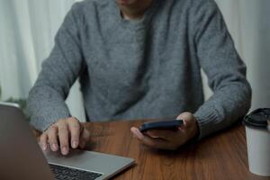 Man in gray sweater using a laptop and holding a smartphone at a wooden table with a coffee cup, focusing on multitasking in a work-from-home setup. photo
