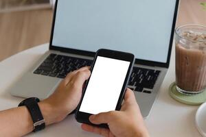 Hands holding blank-screen smartphone in front of laptop on white table, modern remote work setting. photo