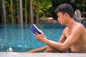 Asian man laying in the swimming pool reading a book in the summer on a sunny day. Relaxing in a backyard swimming pool for relax in summer vacation day. photo