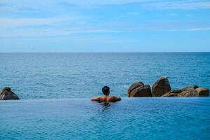 A man relaxing in a swimming pool at a luxury resort with blue sky and blue sea background. Young man enjoying beautiful sunset in luxury hotel near swimming pool. Calmness and serenity. photo