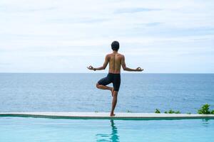 Man doing yoga and meditating in tree position near swimming pool on the tropical beach in Samui, Thailand. Healthy lifestyle active balanced practicing meditate and energy, yoga in morning. photo