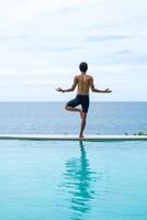 Man doing yoga and meditating in tree position near swimming pool on the tropical beach in Samui, Thailand. Healthy lifestyle active balanced practicing meditate and energy, yoga in morning. photo