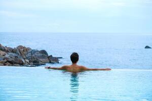 A man relaxing in a swimming pool at a luxury resort with blue sky and blue sea background. Young man enjoying beautiful sunset in luxury hotel near swimming pool. Calmness and serenity. photo