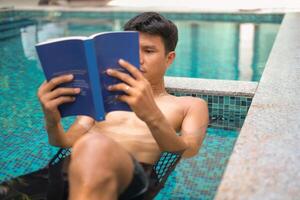 Asian man laying in the swimming pool reading a book in the summer on a sunny day. Relaxing in a backyard swimming pool for relax in summer vacation day. photo