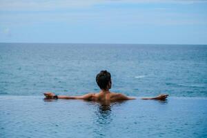 A man relaxing in a swimming pool at a luxury resort with blue sky and blue sea background. Young man enjoying beautiful sunset in luxury hotel near swimming pool. Calmness and serenity. photo