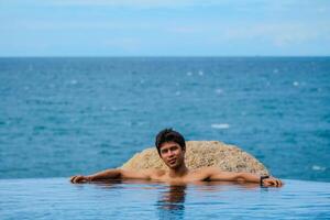 A man relaxing in a swimming pool at a luxury resort with blue sky and blue sea background. Young man enjoying beautiful sunset in luxury hotel near swimming pool. Calmness and serenity. photo