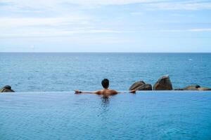 A man relaxing in a swimming pool at a luxury resort with blue sky and blue sea background. Young man enjoying beautiful sunset in luxury hotel near swimming pool. Calmness and serenity. photo