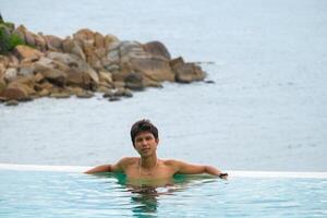 A man relaxing in a swimming pool at a luxury resort with blue sky and blue sea background. Young man enjoying beautiful sunset in luxury hotel near swimming pool. Calmness and serenity. photo