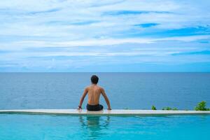 A man relaxing in a swimming pool at a luxury resort with blue sky and blue sea background. Young man enjoying beautiful sunset in luxury hotel near swimming pool. Calmness and serenity. photo