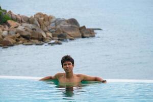 A man relaxing in a swimming pool at a luxury resort with blue sky and blue sea background. Young man enjoying beautiful sunset in luxury hotel near swimming pool. Calmness and serenity. photo