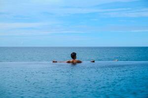 A man relaxing in a swimming pool at a luxury resort with blue sky and blue sea background. Young man enjoying beautiful sunset in luxury hotel near swimming pool. Calmness and serenity. photo