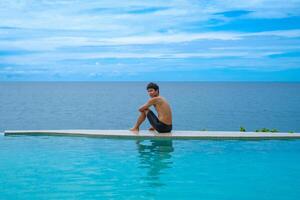 A man relaxing in a swimming pool at a luxury resort with blue sky and blue sea background. Young man enjoying beautiful sunset in luxury hotel near swimming pool. Calmness and serenity. photo