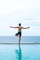 Man doing yoga and meditating in tree position near swimming pool on the tropical beach in Samui, Thailand. Healthy lifestyle active balanced practicing meditate and energy, yoga in morning. photo