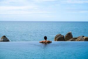 A man relaxing in a swimming pool at a luxury resort with blue sky and blue sea background. Young man enjoying beautiful sunset in luxury hotel near swimming pool. Calmness and serenity. photo
