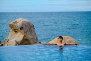 A man relaxing in a swimming pool at a luxury resort with blue sky and blue sea background. Young man enjoying beautiful sunset in luxury hotel near swimming pool. Calmness and serenity. photo