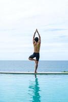 Man doing yoga and meditating in tree position near swimming pool on the tropical beach in Samui, Thailand. Healthy lifestyle active balanced practicing meditate and energy, yoga in morning. photo