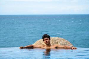 A man relaxing in a swimming pool at a luxury resort with blue sky and blue sea background. Young man enjoying beautiful sunset in luxury hotel near swimming pool. Calmness and serenity. photo