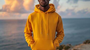 A man in a yellow hoodie stands on a beach with the ocean in the background photo