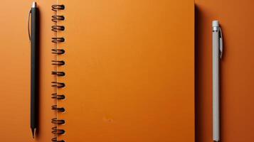 A black pen and a silver pen are on top of an orange notebook photo
