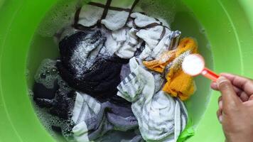 Carefully hand adding laundry detergent to a green bucket filled with various clothes items video