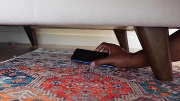 A hand is reaching under the couch in an attempt to retrieve an item that is on a colorful rug video