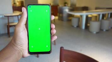 A person is holding a smartphone featuring a green screen while sitting in a cafe setting video
