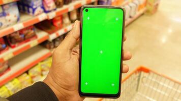 A person is holding a smartphone that features a green screen while in a grocery store setting video