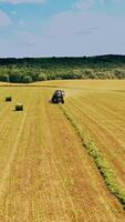 Tractor at work and some bales on the field on natural background. Agricultural tractor in field pressing grass into round bundles at sunny summer day. Aerial view. Vertical video