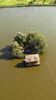 Small island with green trees and a house in the river. Aerial view of island and wooden house in the middle of a lake surrounded by water at daytime in summer. Camera moves down. Vertical video