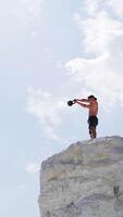 View from below on a healthy man training with heavy weight on a hill. Bodybuilder lifting kettlebell on the natural sky background. Shirtless man during workout outdoors. Vertical video