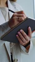 Woman in light jacket standing at the big window. Lady holding a notebook, writing something down there and closing it. Close up. Vertical video
